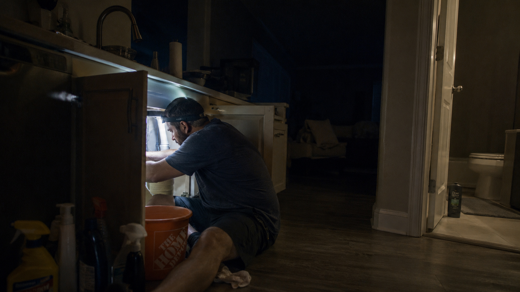 A father fixes the plumbing under the sink at night by headlamp.